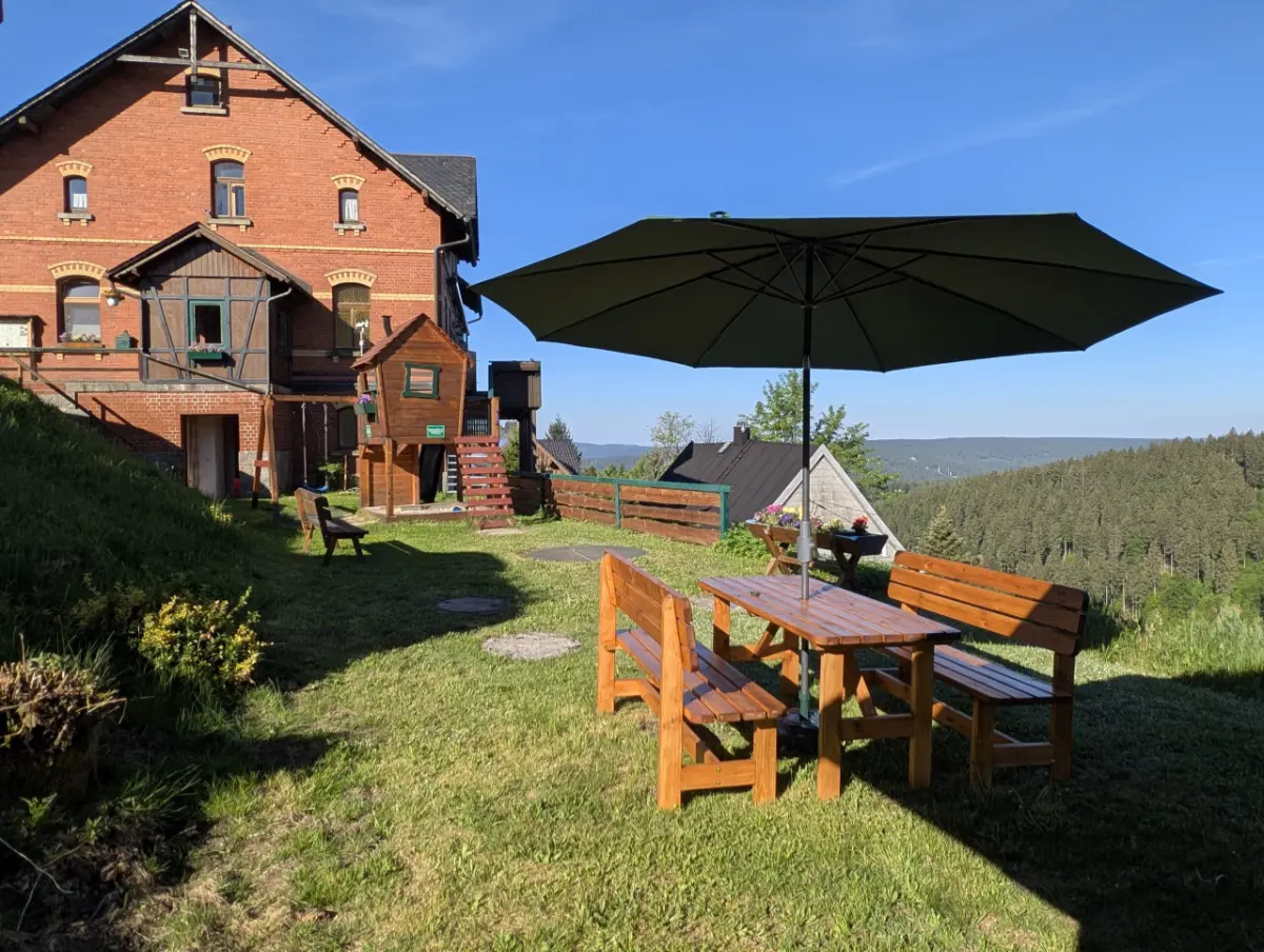 Außenbereich des Gasthaus Schöne Aussicht in Klingenthal mit Sitzgruppe unter einem Sonnenschirm auf einer Wiese, einem Kinderspielhaus und einem großen Backsteingebäude im Hintergrund unter blauem Himmel