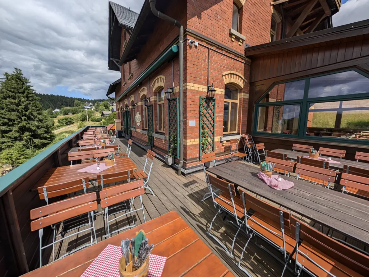 Holzterrasse des Berggasthaus Schöne Aussicht mit zahlreichen leeren Holztischen und Stühlen, gedeckt mit karierten Tischdecken, vor einer Backsteinfassade mit Blick auf ein bewaldetes Tal