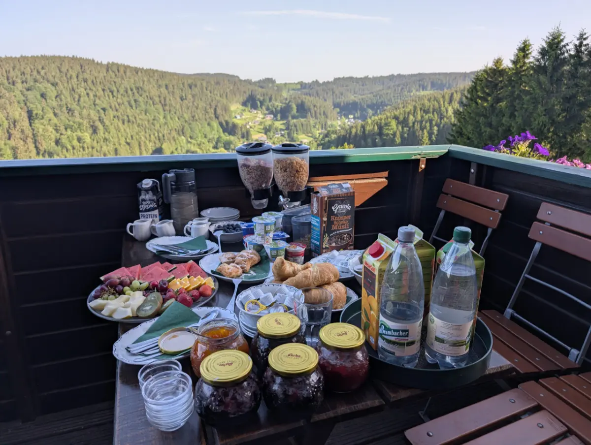 Ein reichhaltiges Frühstücksbuffet auf einem Holztisch auf einem Balkon, mit Obstplatten, Croissants, Müsli-Spendern, Saftflaschen und Marmeladengläsern, vor einem weiten Panoramablick auf bewaldete Hügel