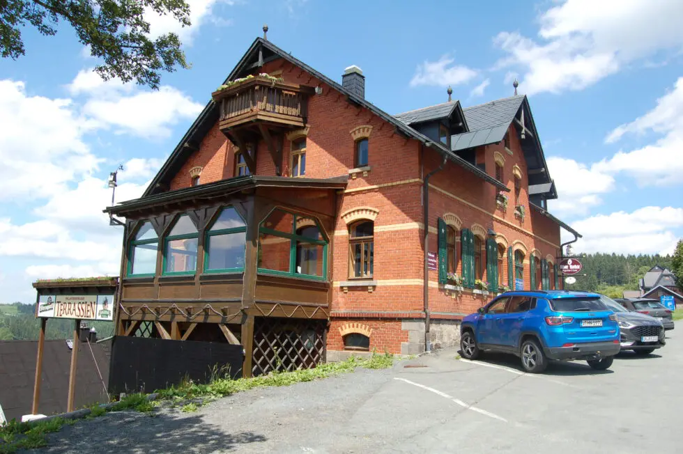 Außenansicht des Gasthauses 'Berggasthaus Schöne Aussicht' in einem historischen, zweigeschossigen Backsteingebäude mit großem Holzvorbau (Veranda), Balkon und Terrasse. Ein blauer SUV und ein weiteres Auto parken auf dem Parkplatz davor, im Hintergrund ist eine Hügellandschaft zu sehen.