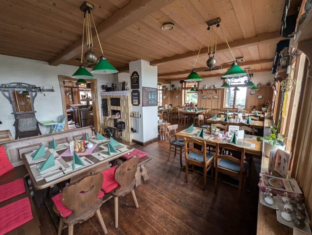 Gaststube Innenbereich Gemütliche, traditionelle Gaststube mit Holzdecke, Dielenboden und eingedeckten Holztischen mit grünen Servietten. Im Raum hängen grüne Zugpendelleuchten über den Tischen, im Hintergrund ist ein Kachelofen sowie eine Durchreiche zum Thekenbereich zu sehen.