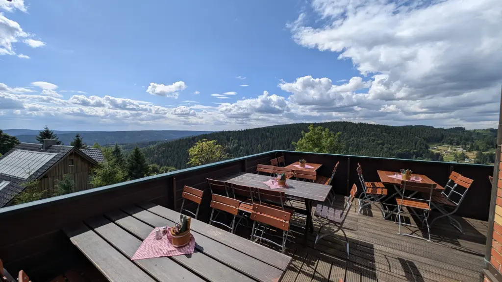 Panoramaterrasse Klingenthal Sonnige Panoramaterrasse mit Holzdielen, gedeckten Tischen und Biergartenstühlen. Im Hintergrund eröffnet sich ein weiter Panoramablick über eine grüne, bewaldete Mittelgebirgslandschaft unter leicht bewölktem Sommerhimmel.