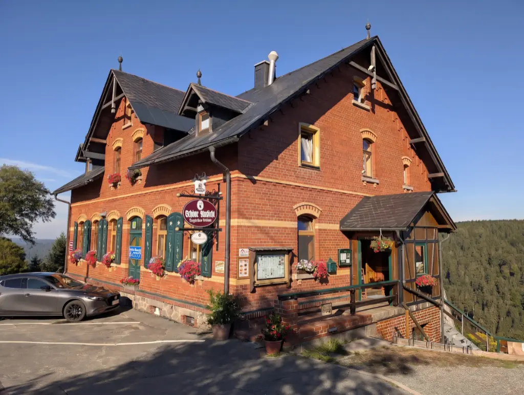 Außenansicht des Gasthauses 'Berggasthaus Schöne Aussicht' in einem historischen, zweigeschossigen Backsteingebäude. Im Hintergrund ist eine Hügellandschaft zu sehen.