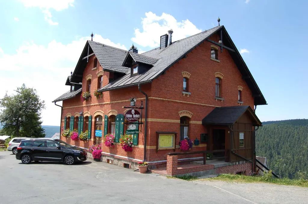 Außenansicht des Berggasthauses Schöne Aussicht: Traditionelles Gebäude aus rotem Backstein mit grün lackierten Fensterläden, reichlich Blumenschmuck an den Fenstern, dunklem Satteldach und einem Parkplatz davor, vor der Kulisse bewaldeter Berge unter blauem Himmel.