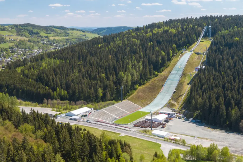 Sparkassen Vogtland Arena Luftaufnahme einer großen Skisprungschanze im Sommer, der Sparkassen Vogtland Arena, die in einen bewaldeten Berghang gebaut ist. Im Vordergrund sind die Zuschauerränge, der Auslaufbereich, Versorgungsgebäude und eine Straße zu sehen.
