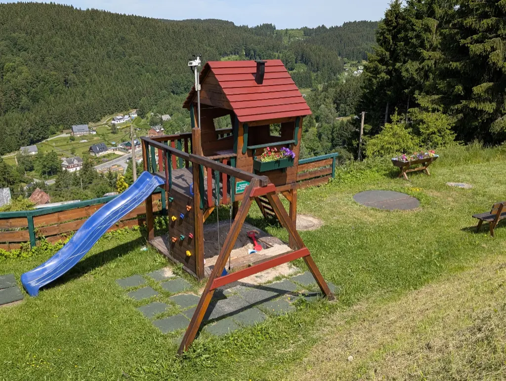 Spielturm für Kinder mit Schaukel und Sandkasten Kinderspielplatz-Set aus Holz mit Rutsche und Sandkasten auf einem Hang im Erzgebirge, mit Blick auf ein Waldtal und Dorf.