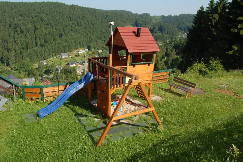Kinderspielplatz-Set aus Holz mit Rutsche und Sandkasten auf einem Hang im Erzgebirge, mit Blick auf ein Waldtal und Dorf.
