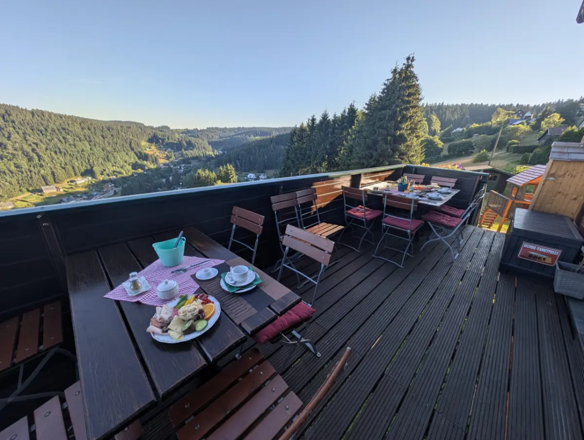 Eine hölzerne Eckterrasse mit zwei gedeckten Tischen und Stühlen mit weinroten Polstern, die einen weiten Panoramablick über ein dicht bewaldetes Tal und ein Dorf im warmen Morgenlicht bietet