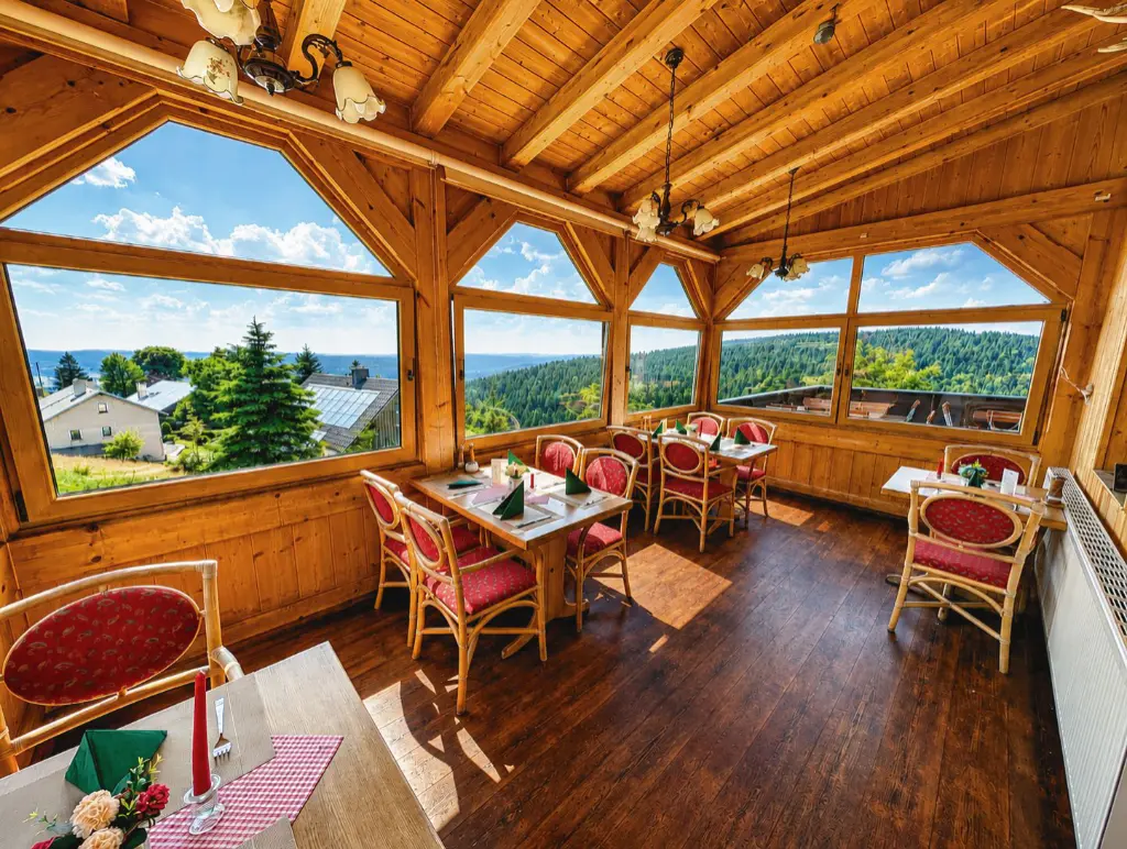Veranda Berggasthaus Schöne Aussicht Ein rustikaler, holzgetäfelter Gastraum in einem Berghotel oder Restaurant. Im Vordergrund sind Tische mit rot gepolsterten Rattanstühlen gedeckt. Die große Fensterfront bietet einen weiten Ausblick auf eine dicht bewaldete Mittelgebirgslandschaft unter blauem Himmel. Kronleuchter hängen von der Holzdecke.