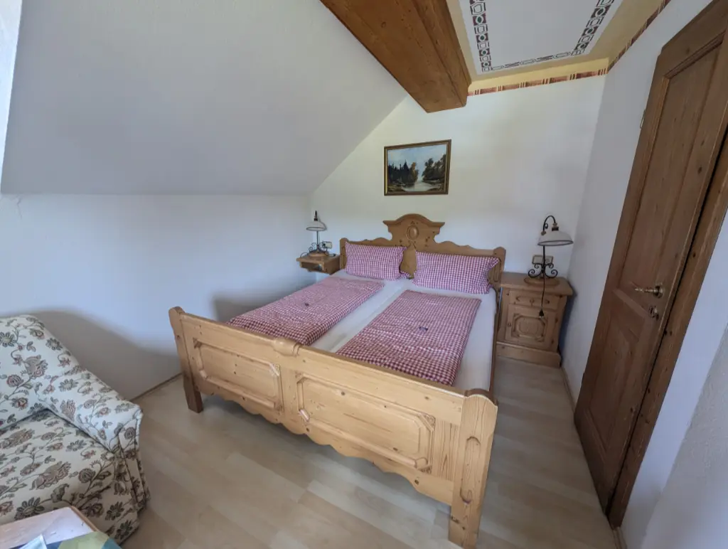 Gemütliches Hotelzimmer im Landhausstil unter einer Dachschräge mit einem massiven Doppelbett aus Holz, bezogen mit rot-weiß karierter Bettwäsche, zwei passenden Nachttischen und einem floral gemusterten Polstersessel im Vordergrund.