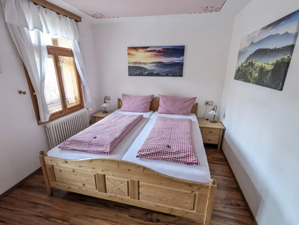 Helles Pensionszimmer im rustikalen Landhausstil mit Holzboden, einem massiven Holz-Doppelbett mit rot-weiß karierter Bettwäsche, zwei Nachttischen mit Tischlampen und Landschaftsbildern an den weißen Wänden.