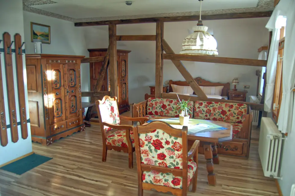 Gemütlicher Wohn- und Schlafbereich in einem rustikalen Fachwerkhaus-Apartment. Im Vordergrund eine gemütliche Sitzecke mit geschnitztem Holztisch, einem Sofa und Sesseln mit rotem Rosenmuster. Links ein prachtvoller, handbemalter Holzschrank. Durch die sichtbaren Deckenbalken hindurch ist im Hintergrund das Doppelbett unter einer Dachschräge zu sehen. Das Fenster auf der rechten Seite zeigt die blaue Stunde, während warmes Licht der Deckenlampe den Raum erfüllt.