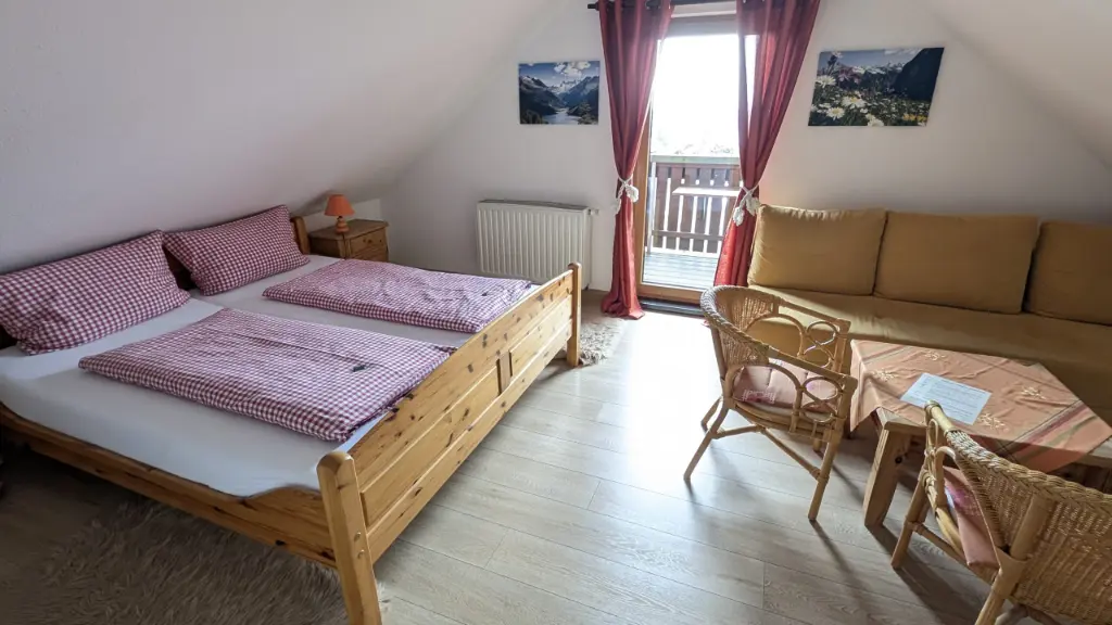 Helles Dachgeschoss-Zimmer im Landhausstil mit einem massiven Holz-Doppelbett und rot-weiß karierter Bettwäsche. Daneben befindet sich ein gemütlicher Wohnbereich mit beigem Sofa, Couchtisch und Rattansesseln sowie eine Balkontür mit roten Vorhängen und Bergbildern an der Wand.