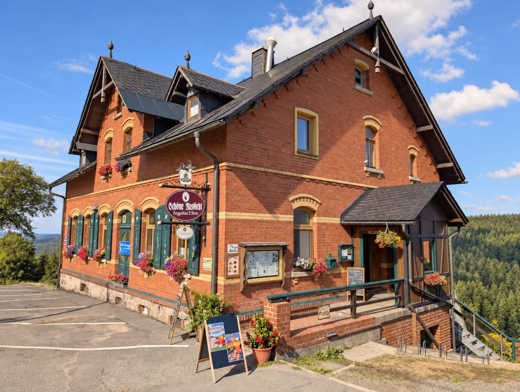 Außenansicht des Gasthauses 'Berggasthaus Schöne Aussicht' in einem historischen, zweigeschossigen Backsteingebäude. Im Hintergrund ist eine Hügellandschaft zu sehen.