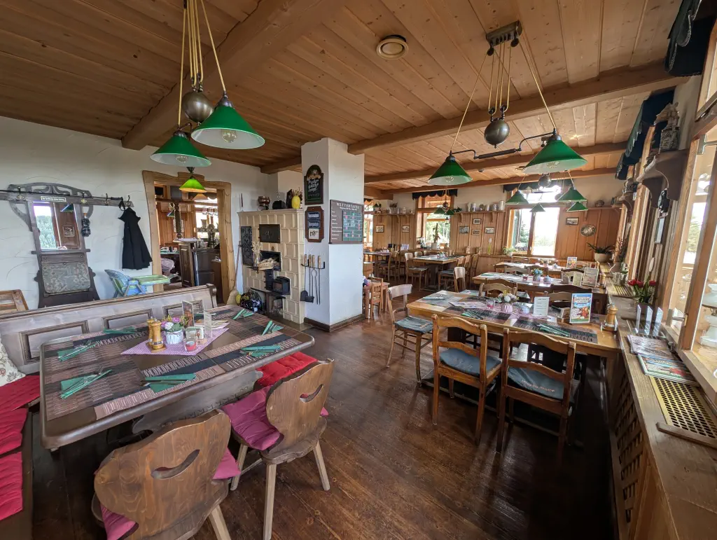 Gemütliche, traditionelle Gaststube mit Holzdecke, Dielenboden und eingedeckten Holztischen mit grünen Servietten. Im Raum hängen grüne Zugpendelleuchten über den Tischen, im Hintergrund ist ein Kachelofen sowie eine Durchreiche zum Thekenbereich zu sehen.