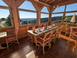 Ein rustikaler, holzgetäfelter Gastraum in einem Berghotel oder Restaurant. Im Vordergrund sind Tische mit rot gepolsterten Rattanstühlen gedeckt. Die große Fensterfront bietet einen weiten Ausblick auf eine dicht bewaldete Mittelgebirgslandschaft unter blauem Himmel. Kronleuchter hängen von der Holzdecke.