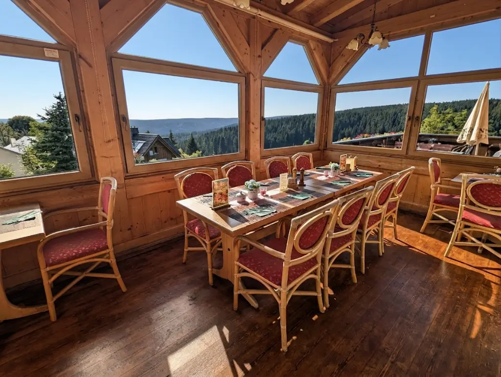 Veranda Ein rustikaler, holzgetäfelter Gastraum in einem Berghotel oder Restaurant. Im Vordergrund sind Tische mit rot gepolsterten Rattanstühlen gedeckt. Die große Fensterfront bietet einen weiten Ausblick auf eine dicht bewaldete Mittelgebirgslandschaft unter blauem Himmel. Kronleuchter hängen von der Holzdecke.
