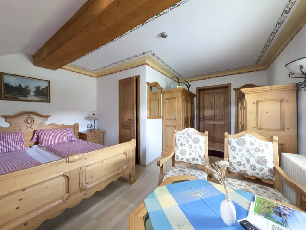 Helles Dachgeschoss-Zimmer im Landhausstil mit einem massiven Holz-Doppelbett und rot-weiß karierter Bettwäsche. Daneben befindet sich ein gemütlicher Wohnbereich mit beigem Sofa, Couchtisch und Rattansesseln sowie eine Balkontür mit roten Vorhängen und Bergbildern an der Wand.