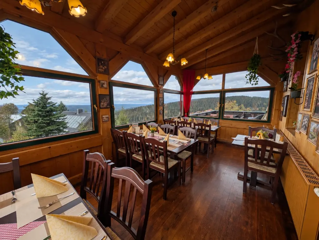 Ein rustikaler, holzgetäfelter Gastraum in einem Berghotel oder Restaurant. Im Vordergrund sind Tische mit rot gepolsterten Rattanstühlen gedeckt. Die große Fensterfront bietet einen weiten Ausblick auf eine dicht bewaldete Mittelgebirgslandschaft unter blauem Himmel. Kronleuchter hängen von der Holzdecke.